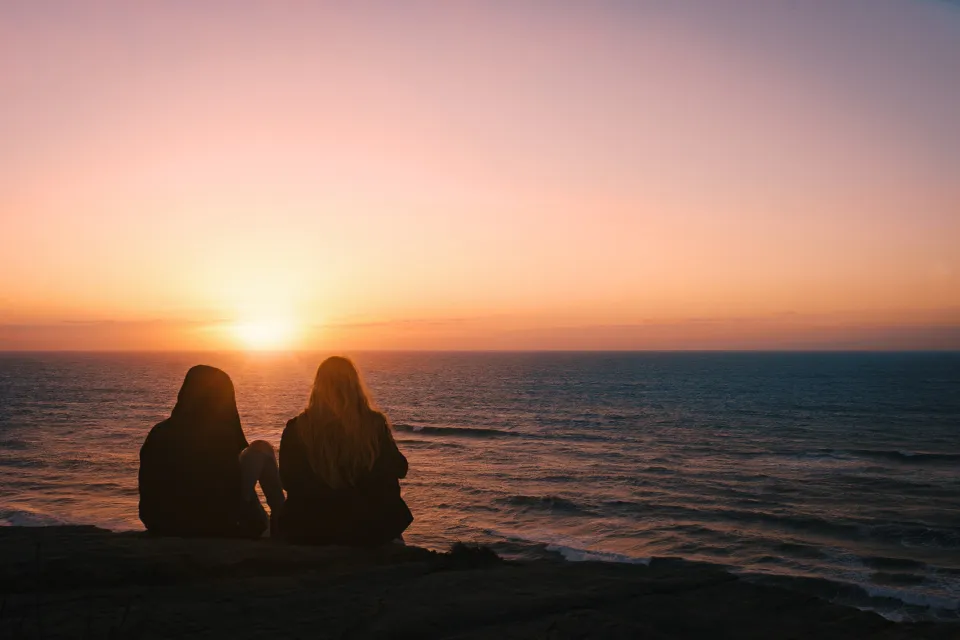 Two people sitting by the ocean at sunset, reflecting on the cost of a basic cremation in Australia